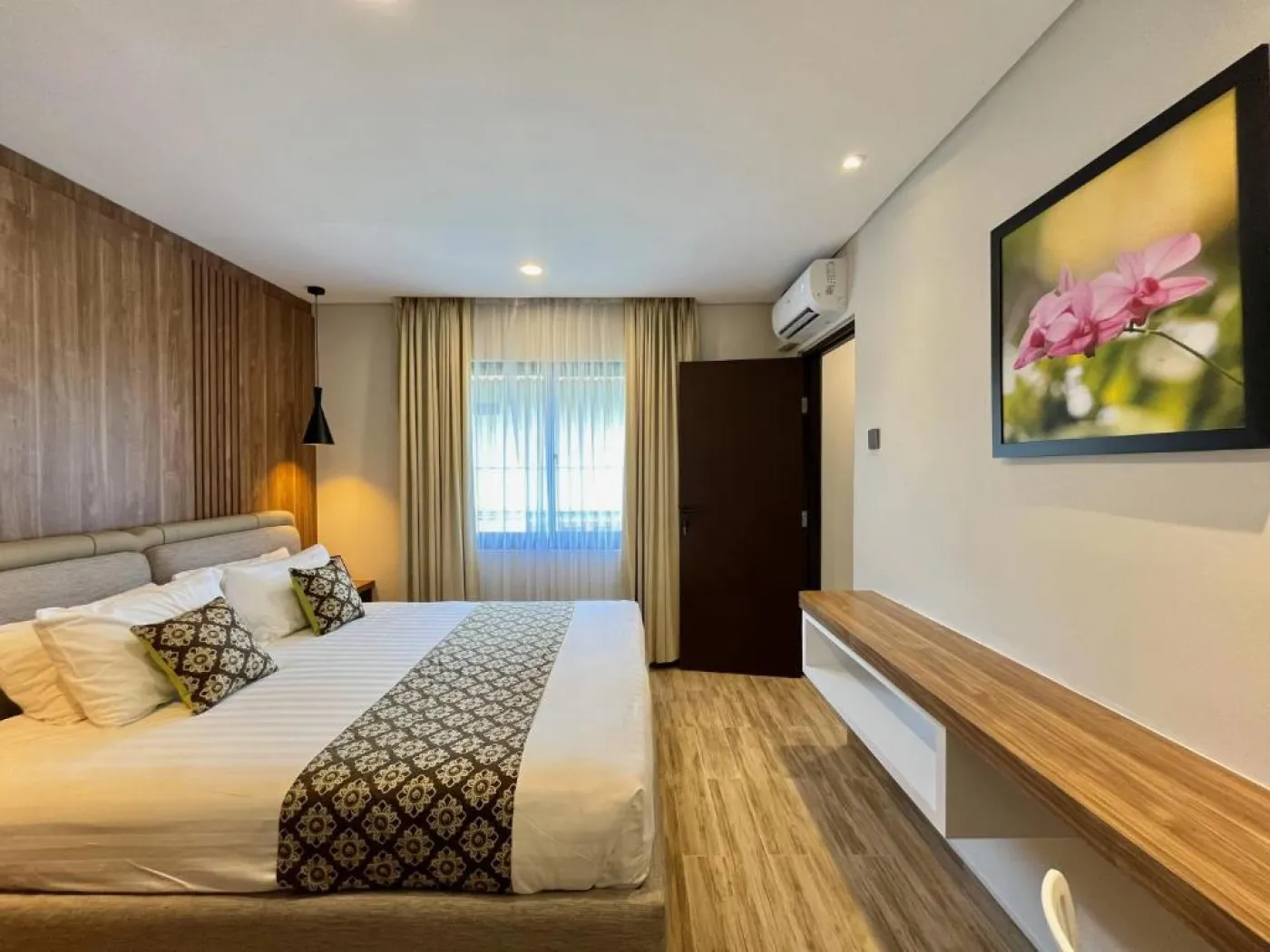 Modern hotel bedroom featuring a king-sized bed with patterned linens, wooden accent wall, floral artwork, and large window with sheer curtains. - Bali Villas